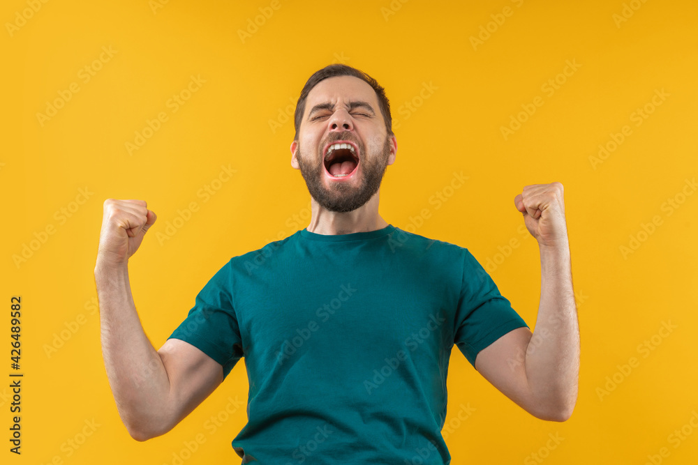 Studio portrait of joyful, excited, glad bearded man with wide open mouth and clenched fists celebrating money win in online lottery or at bookmaker's, isolated over yellow background