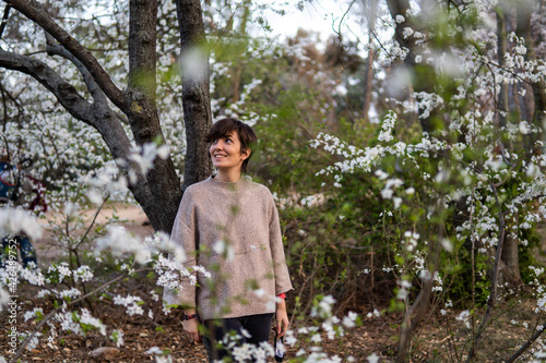 Photo of a young and attractive girl in the park surrounded by almond trees during spring. She's wearing a jumper and smiling