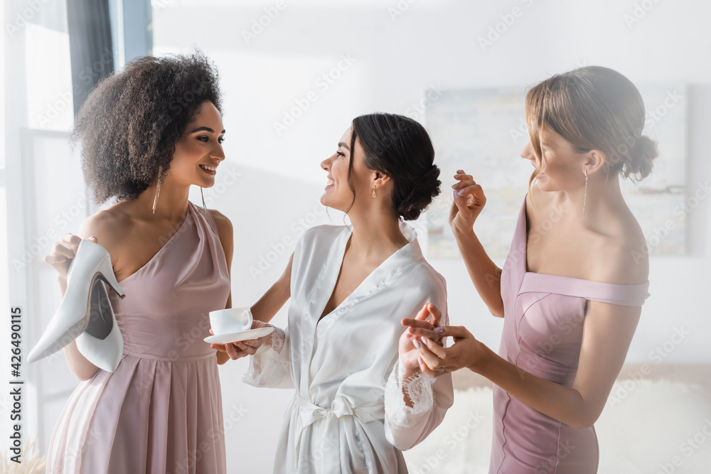 Fototapeta premium cheerful woman in silk robe smiling near interracial friends preparing her for wedding.