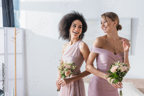 joyful multicultural bridesmaids holding wedding bouquets in bedroom. photo