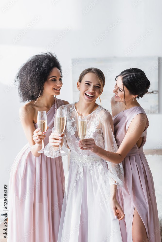 laughing bride holding champagne near cheerful interracial friends in bedroom. laughing bride holding champagne near cheerful interracial friends in bedroom.