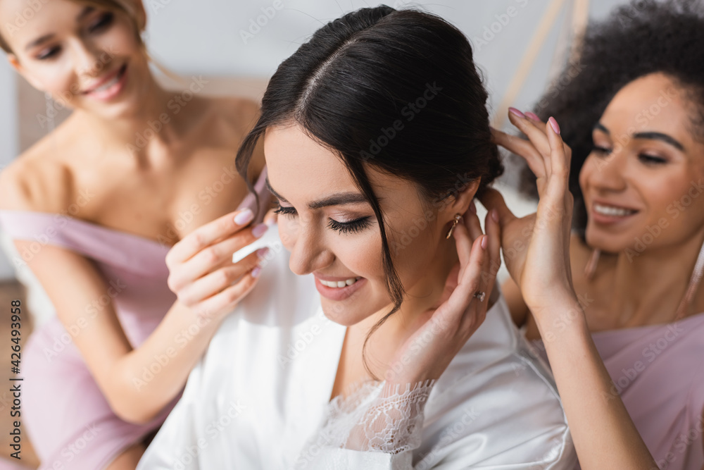 interracial bridesmaids doing hairstyle to happy bride, blurred background.