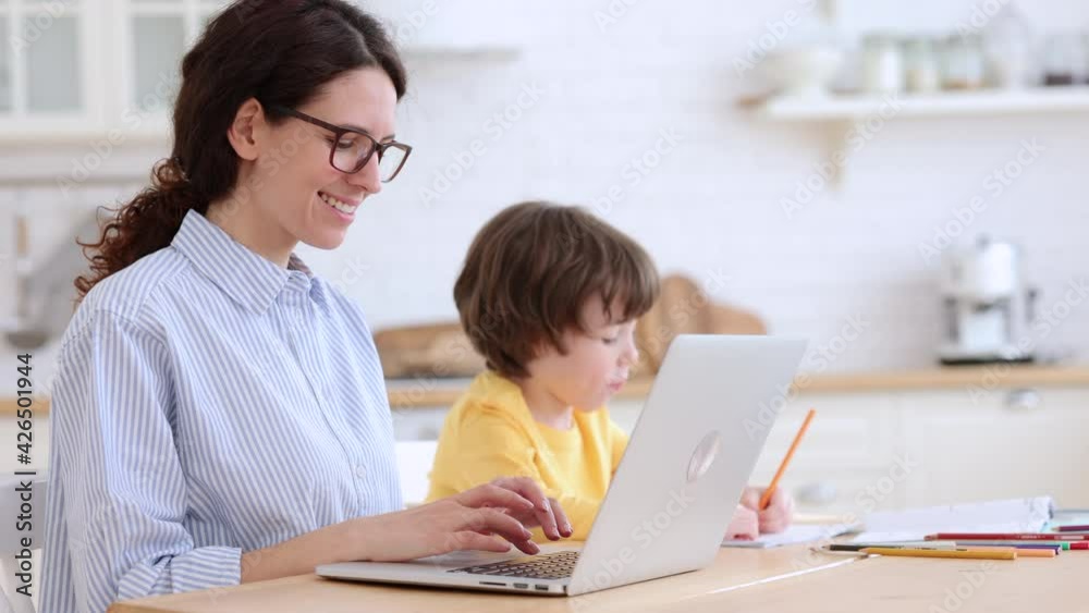 Family, lockdown distance job concept. Kid son painting while his mother is working on laptop at home office. Woman with her child doing homework or studying sitting together at table in living room. Family, lockdown distance job concept. Kid son painting while his mother is working on laptop at home office. Woman with her child doing homework or studying sitting together at table in living room.