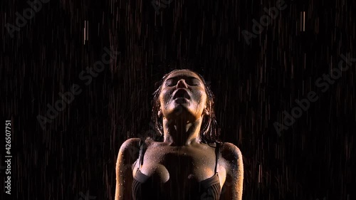 Sensual woman creates splashes of hair and strokes body with hands. Beautiful sexy brunette posing in rain. Silhouette of young sexy female body in underwear in drops of water. Close up. Slow motion.