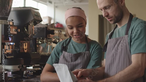 Tilt up of middle-aged Caucasian male barista standing at worktop in cafe and explaining new female mixed-race colleague how to choose necessary coffee typeTilt up of middle-aged Caucasian male barist photo