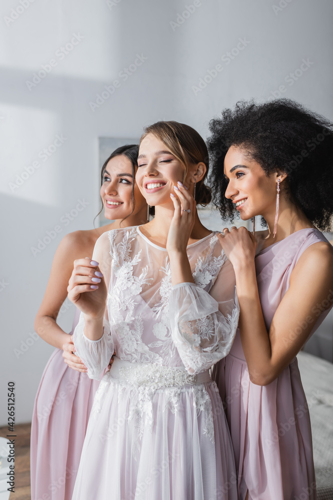 happy bride with closed eyes touching face near smiling interracial bridesmaids. happy bride with closed eyes touching face near smiling interracial bridesmaids.