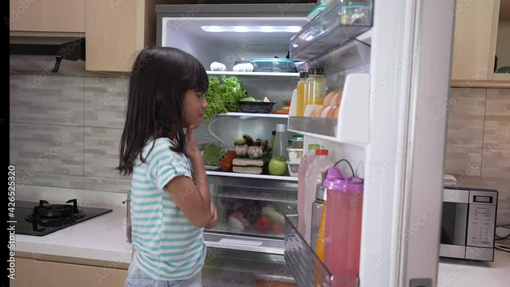 happy young asian girl open fridge door looking for food and eating fruit happy young asian girl open fridge door looking for food and eating fruit