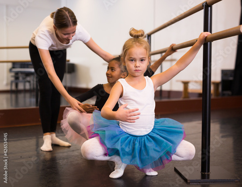 Young female classical dance teacher helping her little students near ballet barre in dancing hall