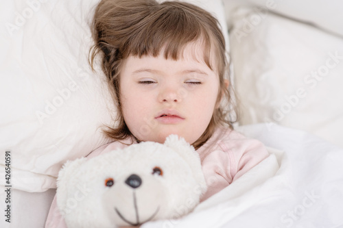 A girl with down syndrome lying on the bed under the covers and hugging a teddy bear. Usually childhood in a family for children with disabilities