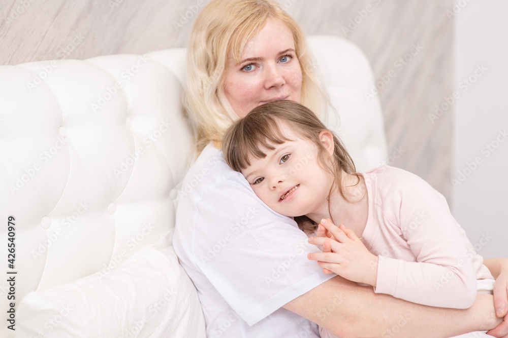 Mom hugging a girl with down syndrome at home in the bedroom on the bed. Ordinary childcare in a family for children with disabilities Mom hugging a girl with down syndrome at home in the bedroom on the bed. Ordinary childcare in a family for children with disabilities