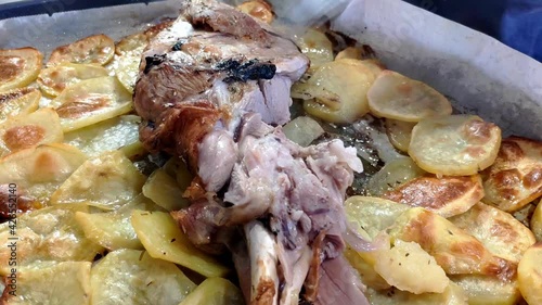 Tray with a roasted lamb shank and potatoes. Pieces are being cut from the lamb and taken. Some potatoes have brown toasted edges. Steam is cominf of the food. photo