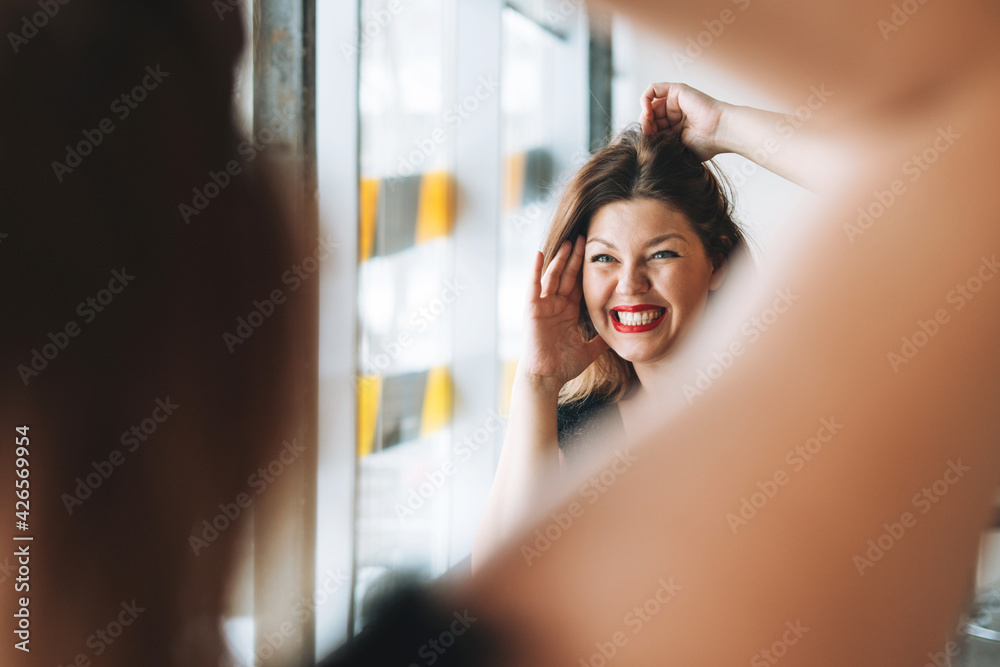 Beautiful smiling young woman plus size body positive in black evening dress make hair style near mirror at the beauty studio Beautiful smiling young woman plus size body positive in black evening dress make hair style near mirror at the beauty studio