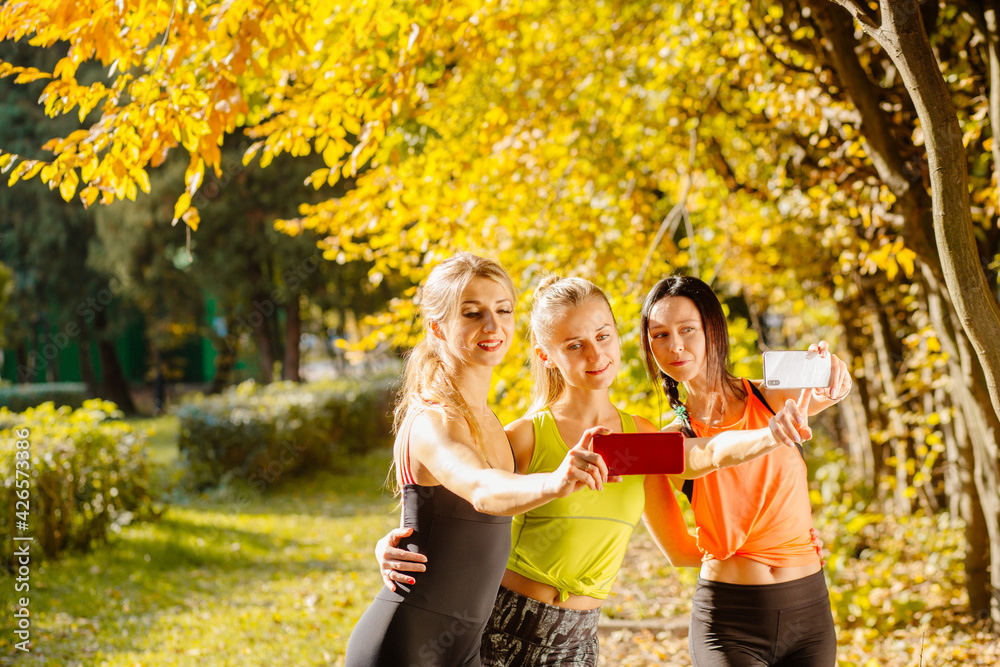 Fototapeta premium Group of women in their 30s working out together in the outdoors. Three females dressed in sportswear taking a selfie on a mobile phone in autumn park, outdoor.