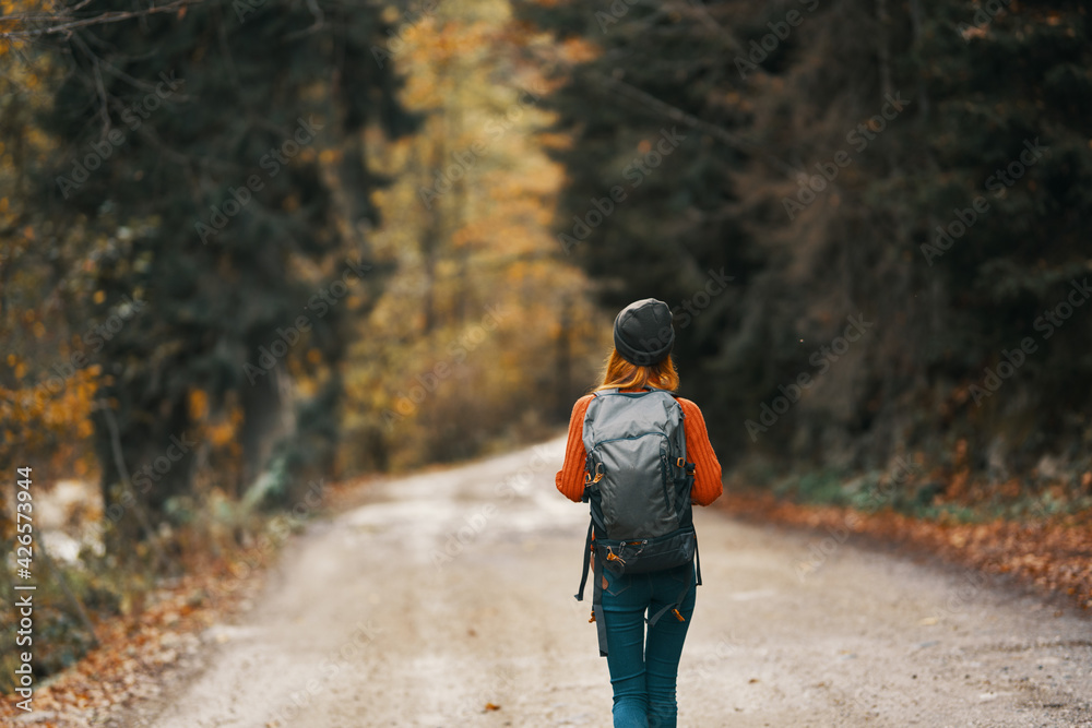 woman with a backpack in a hat and an orange sweater on the road in the autumn forest woman with a backpack in a hat and an orange sweater on the road in the autumn forest