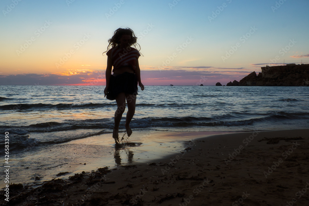 Teenage girl jumping in the sand and water at Golden Bay Beach in Malta on a fall evening at sunset.