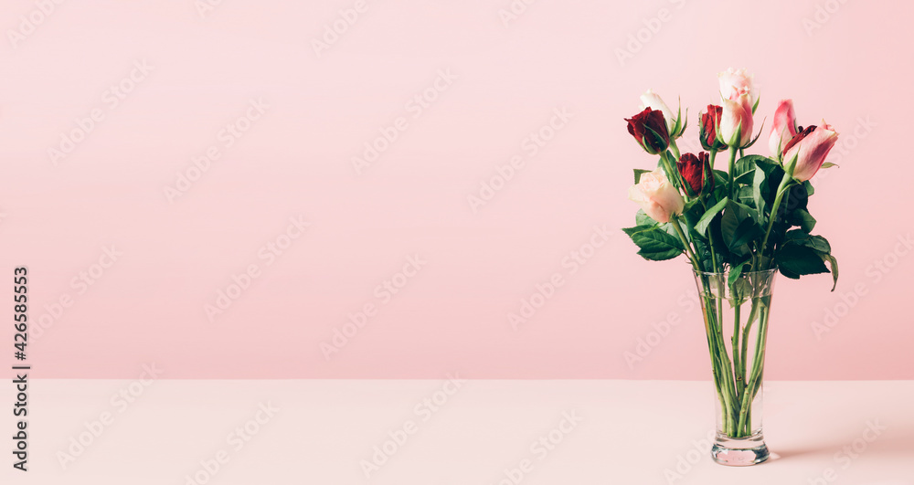 Beautiful fresh roses in a glass vase are arranged on a table against a pastel pink background. Selective focus copy space