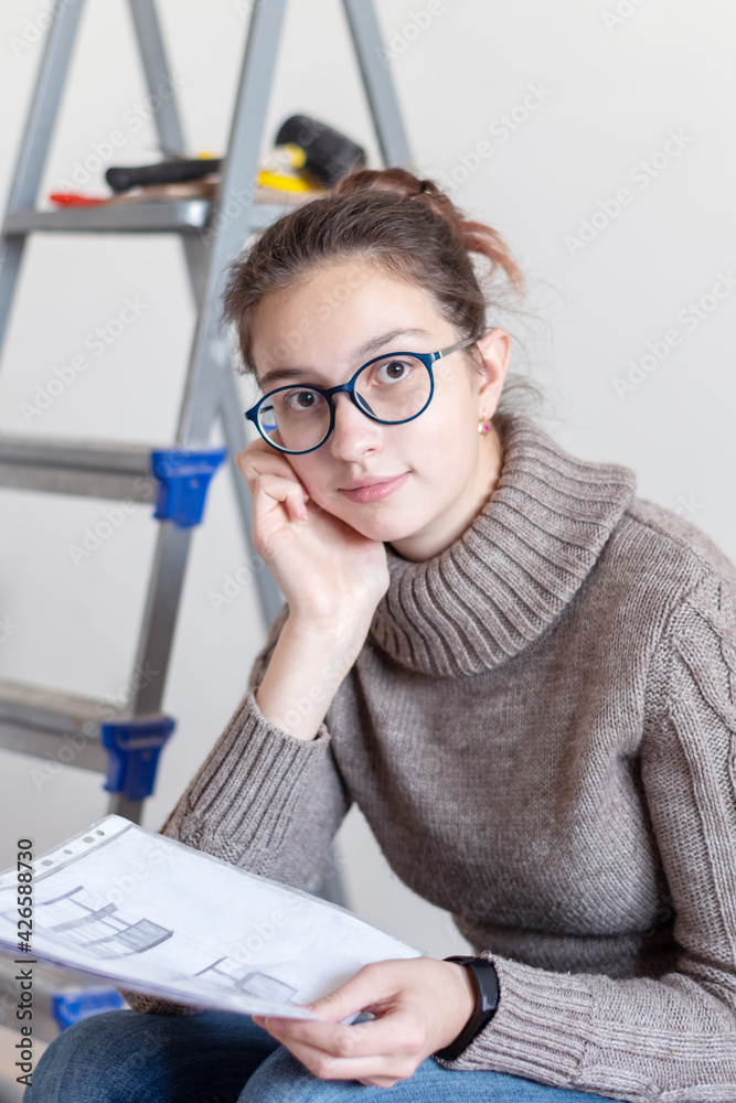 Repair, renovation concept. Caucasian girl designer, architect in blue glasses and a beige sweater holds a blueprint and looks at you at home. Repair, renovation concept. Caucasian girl designer, architect in blue glasses and a beige sweater holds a blueprint and looks at you at home.