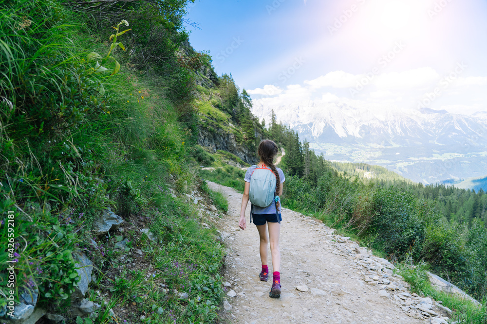 Fototapeta premium Girl hiking on beautiful summer day in alps mountains Austria, resting on rock and admire amazing view to mountain peaks. Active family vacation leisure with kids. Outdoor fun and healthy activity