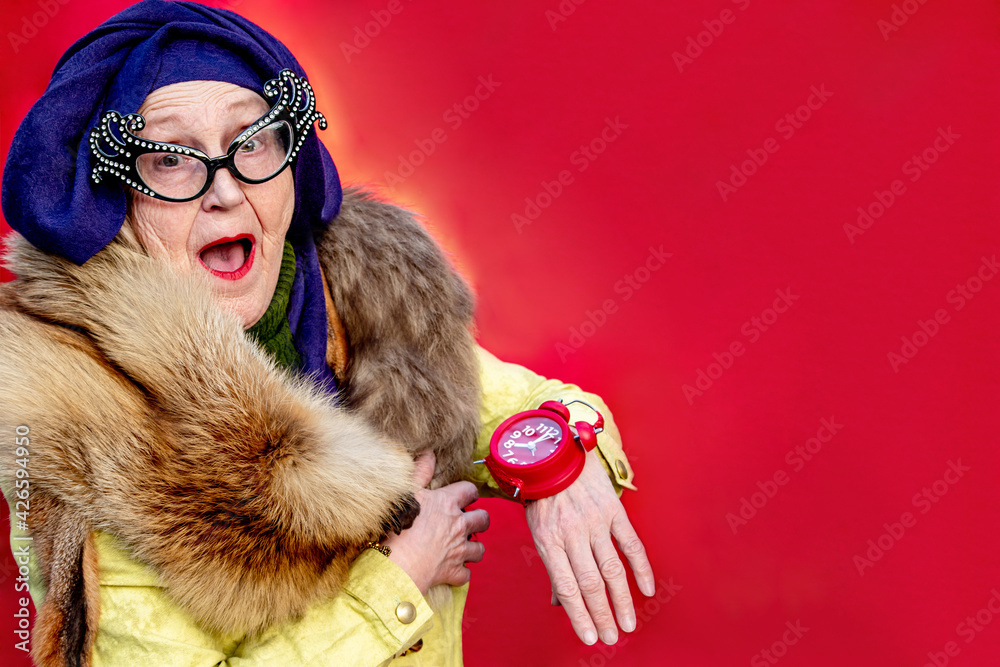 Glamorous Old woman wears an alarm clock like a wristwatch. Stylish older woman in turban and glamorous glasses. Grandma shows watch on her arm. Hurrying cheerful elderly fashionista. Emotional people Glamorous Old woman wears an alarm clock like a wristwatch. Stylish older woman in turban and glamorous glasses. Grandma shows watch on her arm. Hurrying cheerful elderly fashionista. Emotional people