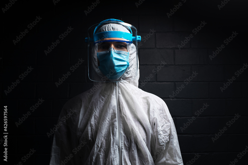 Dark studio portrait of young doctor wearing PPE suit against coronavirus and covid-19. Background of black brick wall. Dark studio portrait of young doctor wearing PPE suit against coronavirus and covid-19. Background of black brick wall.