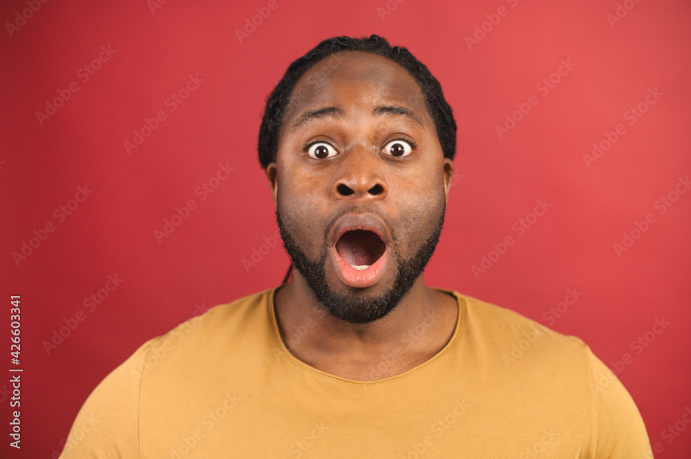 Close portrait of surprised african bearded man with opened mouth isolated on red background, with afro haircut in mustard t-shirt looking in camera with raised eyebrows and surprised face expression Close portrait of surprised african bearded man with opened mouth isolated on red background, with afro haircut in mustard t-shirt looking in camera with raised eyebrows and surprised face expression
