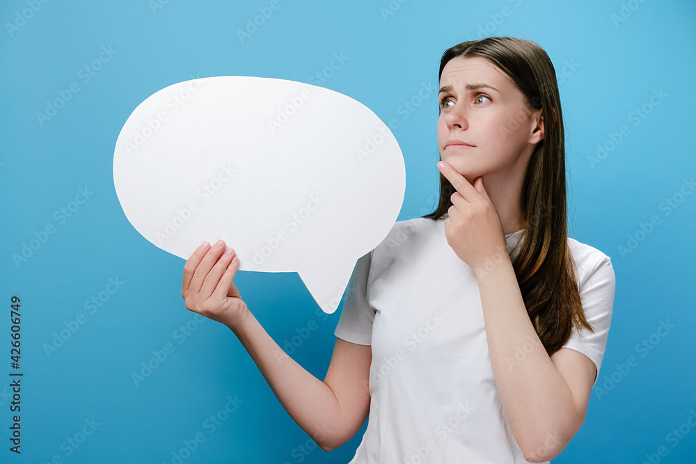 Worried pensive confused young female holding empty speech bubble, spreading arms put hand prop up on chin, dressed in white t-shirt, isolated on blue studio background wall. Emotion people concept Worried pensive confused young female holding empty speech bubble, spreading arms put hand prop up on chin, dressed in white t-shirt, isolated on blue studio background wall. Emotion people concept