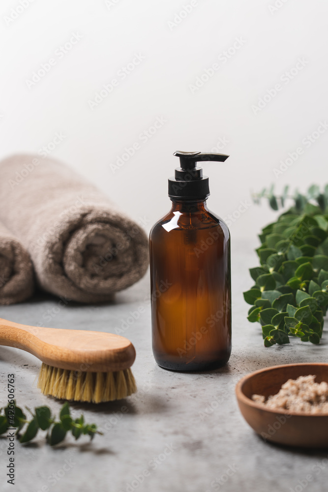 Spa body care. Transparent glass bottle with dispenser, body scrub in wooden bowl, natural massage brush and eucalyptus branches. Side view, close up view Spa body care. Transparent glass bottle with dispenser, body scrub in wooden bowl, natural massage brush and eucalyptus branches. Side view, close up view
