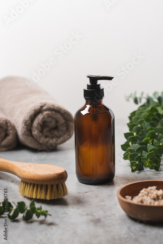 Spa body care. Transparent glass bottle with dispenser, body scrub in wooden bowl, natural massage brush and eucalyptus branches. Side view, close up view