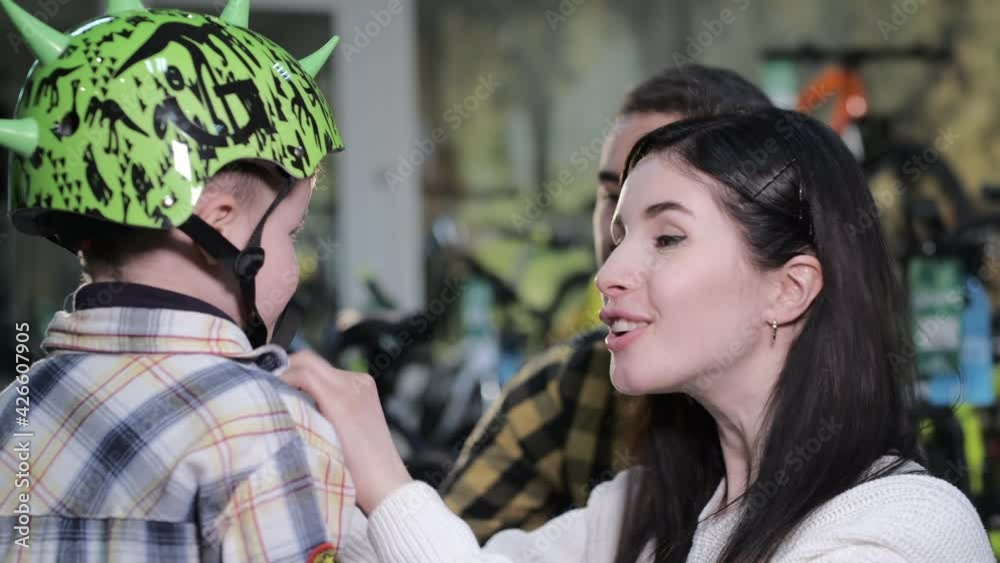 Happy young mother puts on designer helmet to little son head choosing equipment with father in contemporary sports shop closeup