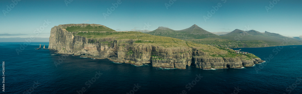 Panoramic view on coastline of island Eysturoy with rock formations Risin og Kellingin on Faroe Islands during the day with sun and blue sky, blue ocean and green cliffs. Panoramic view on coastline of island Eysturoy with rock formations Risin og Kellingin on Faroe Islands during the day with sun and blue sky, blue ocean and green cliffs.