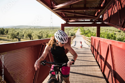 young female cyclist pedals on a red bridge outdoors wearing sunglasses and helmet and checks her smart watch