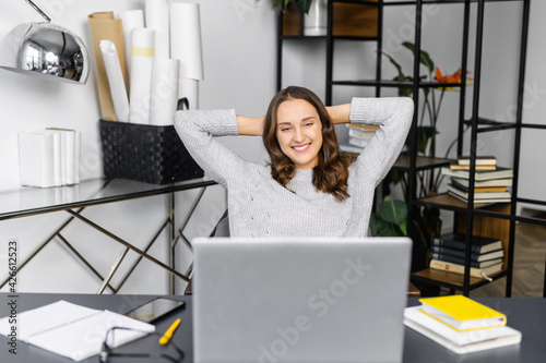 Satisfied young woman leaned back in front of the laptop sitting at the office desk, female employee in casual wear rejoyce finished project, looks at the screen, smiles, takes a break on a workplace photo