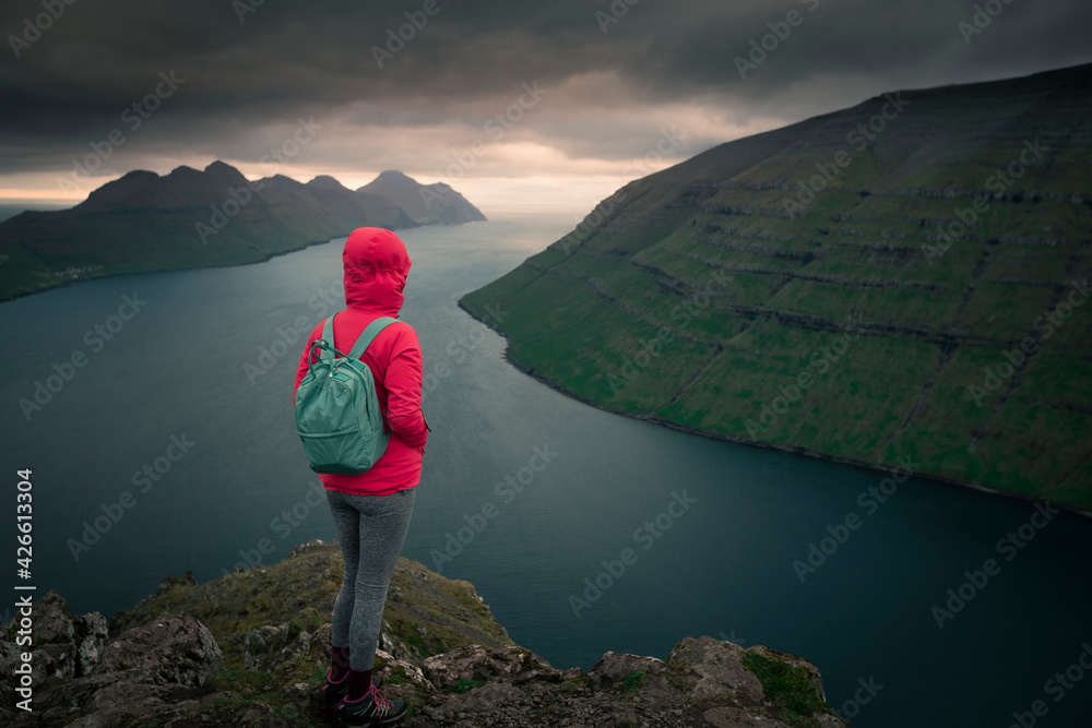 Woman with rain jacket and backpack at the Klakkur viewpoint near Klaksvik on the island of Bordoy with a view of Kalsoy and Kunoy in sunset, Faroe Islands.