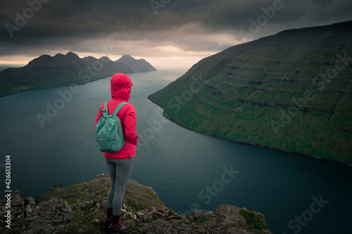 Woman with rain jacket and backpack at the Klakkur viewpoint near Klaksvik on the island of Bordoy with a view of Kalsoy and Kunoy in sunset, Faroe Islands. © Bastian Linder