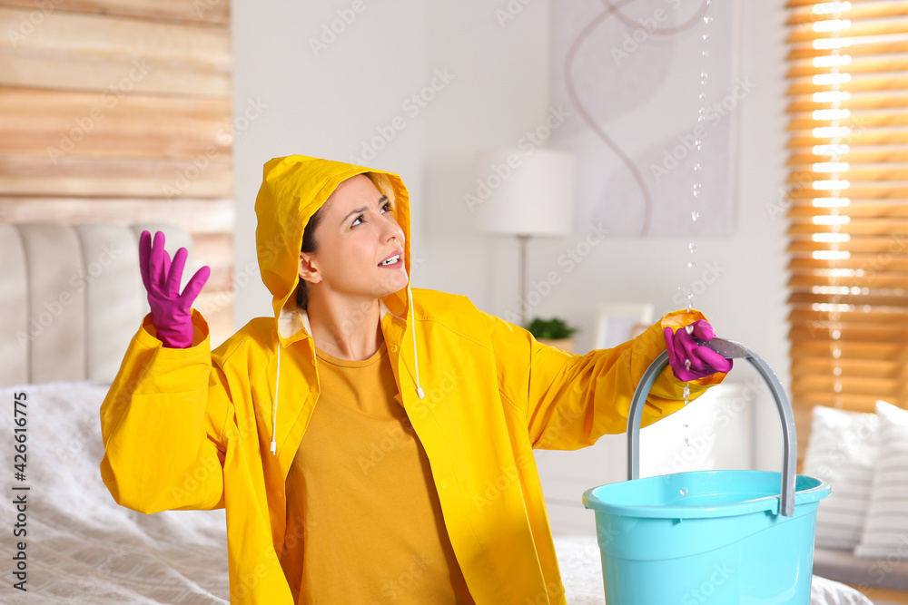 Young woman in raincoat collecting leaking water from ceiling at home. Time to call roof repair service Young woman in raincoat collecting leaking water from ceiling at home. Time to call roof repair service