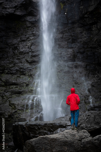 Man stands on rock with red jacket, looks at Fossa waterfall on Streymoy Island, Faroe Islands.