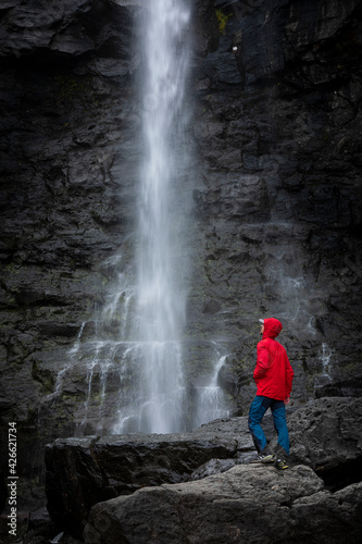 Man stands on rock with red jacket, looks at Fossa waterfall on Streymoy Island, Faroe Islands.