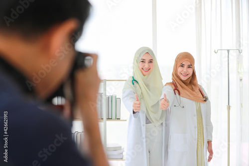 Studio portrait shot of two adult Muslim female doctors with cute smiling, looking at the camera and showing a thumb up gesture. Selective focus at the doctors with blurred cameraman in the foreground