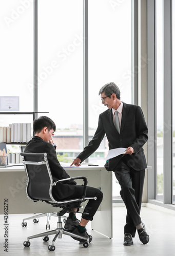 Wide view portrait shot of two Asian businessmen in the office. Senior leader pointing to the paper to show the mistake made by subordinate and blaming him for error while holding a paper document
