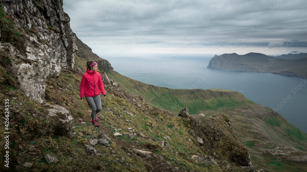 Obraz premium Female hiker with magenta jacket and tights in mountain landscape of Streymoy island with cloudy sky, Faroe Islands.