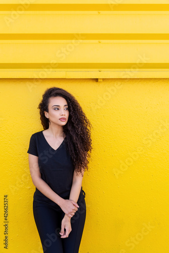 Stylish young black woman with long hair in black outfit in front of a vibrant yellow background