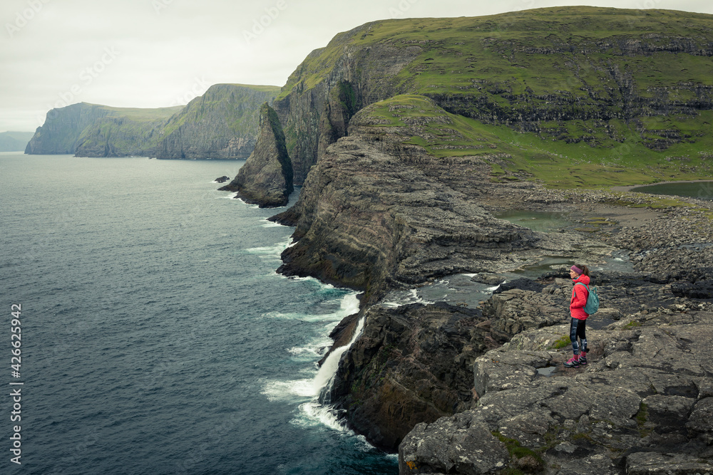 Woman in magenta jacket on coast, waterfall and cliffs at Trælanípa on the island of Vagar, on Lake Leitisvatn, Faroe Islands.