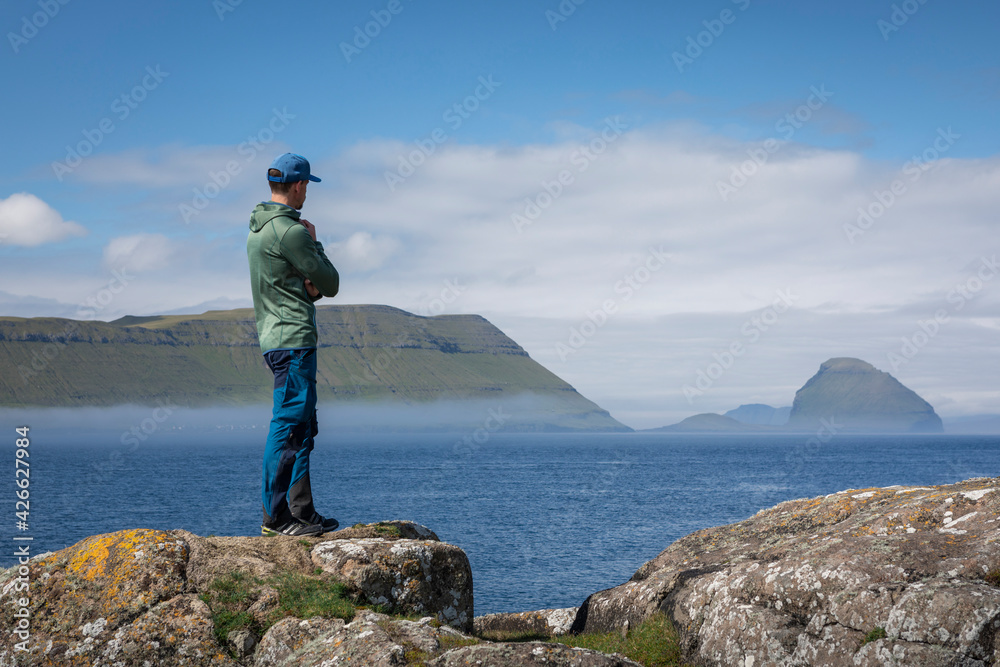 Man on a rock at the coast overlooking islands of the Faroe Islands in sunshine.