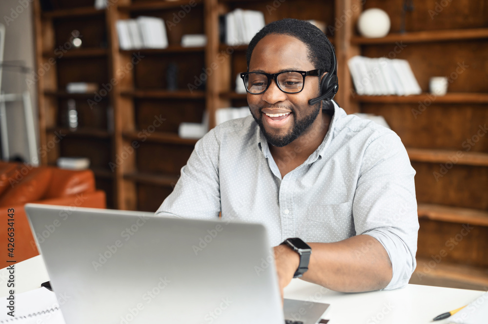 A multiracial guy using a hands free device for online communication sitting at the desk in modern office space, an African-American wearing headset holding video conference, webinar A multiracial guy using a hands free device for online communication sitting at the desk in modern office space, an African-American wearing headset holding video conference, webinar