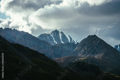 Atmospheric mountain landscape with silhouettes of rocks and snowbound sharp pinnacle under cloudy sky. Great rocks with snow. Beautiful mountain scenery with snow peaked top. Snow-capped pointy peak.