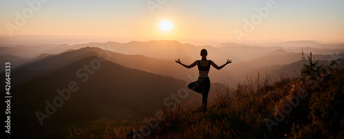 Panoramic view. Back view of slim woman in black sportswear doing yoga exercise on one leg in mountains. Concept of yoga time at sunset.