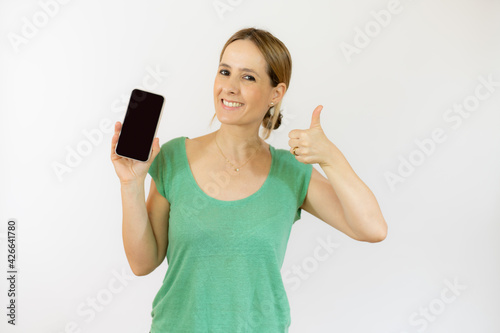 Portrait of a beautiful young woman wearing casual t-shirt with thumb up showing mobile phone isolated over white background