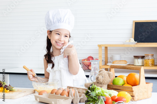 Little Caucasian girl wearing white chef hat happy cooking in kitchen. Child preparing easy food or bakery and lerning to cook by herselfe.