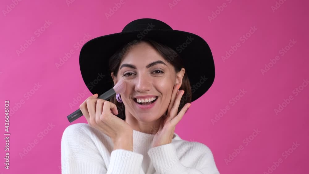 Woman in casual white sweater and hat on pink background isolated happy put blush using brush smile Woman in casual white sweater and hat on pink background isolated happy put blush using brush smile