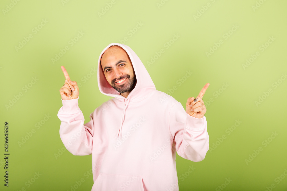 Young bald caucasian man in pink hoodie isolated on green background look to camera with smile point fingers up Young bald caucasian man in pink hoodie isolated on green background look to camera with smile point fingers up